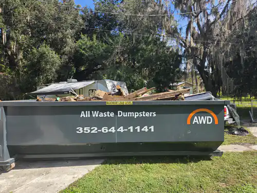 All Waste Dumpsters container loaded level with old patio lumber on the lawn of a Summerfield home, with a green tarp and trash can beside it