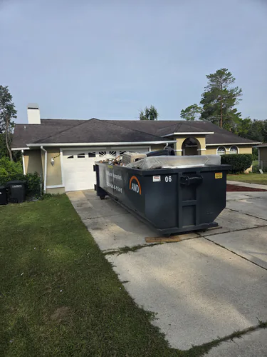 All Waste Dumpsters container number 06 on boards in the concrete driveway of a stucco home, loaded with a mattress and a pet crate