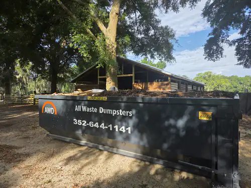 Loaded All Waste Dumpsters container on packed sand under a live oak beside an open wooden barn on a Marion County horse property