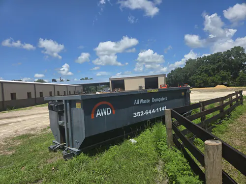 All Waste Dumpsters roll off container on grass beside a dark board fence at a Marion County horse farm with metal barns behind it