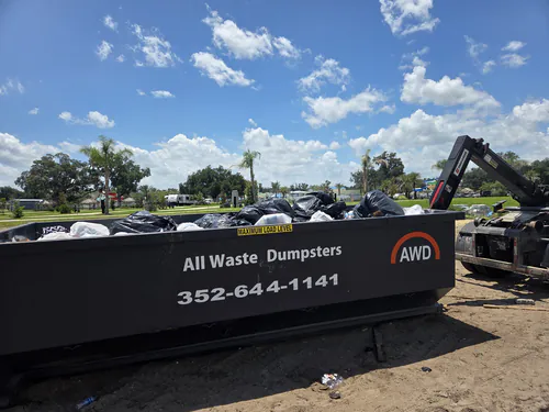 Full All Waste Dumpsters container of black trash bags at an RV resort with palm trees and the truck hook arm raised for pickup