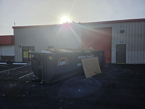 Loaded All Waste Dumpsters container on a dark paved warehouse lot at sunset, with a sheet of plywood leaning against its side