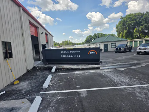 Loaded All Waste Dumpsters container against a warehouse beside an open red roll up door on a freshly paved commercial lot