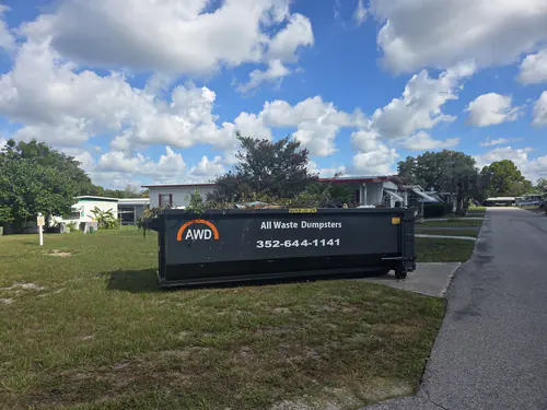 All Waste Dumpsters container loaded with cut brush on the grass beside a narrow paved lane lined with park homes in Ocala