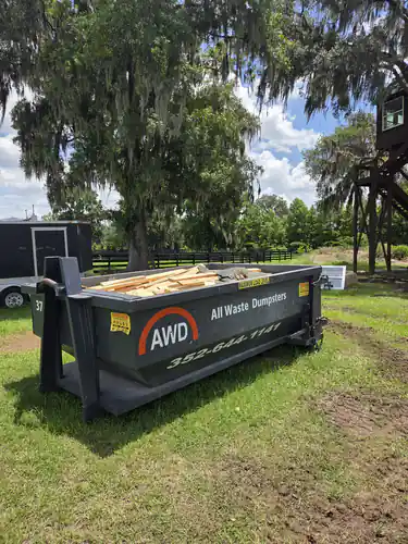 All Waste Dumpsters container number 37 filled with lumber offcuts on grass beside a black board fence and a live oak in Ocala