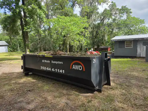 All Waste Dumpsters container number 29 loaded with cut brush on a wooded Ocala lot beside two sheds and a riding mower