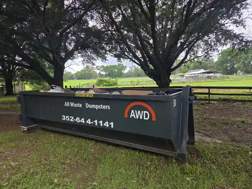 Full All Waste Dumpsters 25 yard container on grass under live oaks with a board fence, pasture and barn behind it on an Ocala property