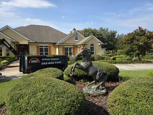 All Waste Dumpsters truck setting a container in the driveway of a yellow stucco home in Summerfield with trimmed shrubs and a rearing horse statue in front