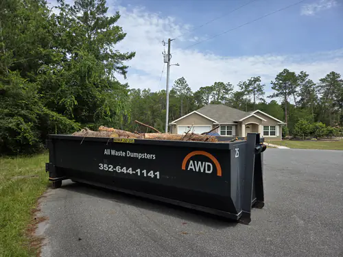 All Waste Dumpsters container number 25 loaded with pine logs and limbs on the asphalt at the edge of a wooded Summerfield street