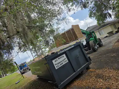 Green telehandler raising a plywood bin beside an All Waste Dumpsters container in the driveway of a yellow home under oaks in Marion County